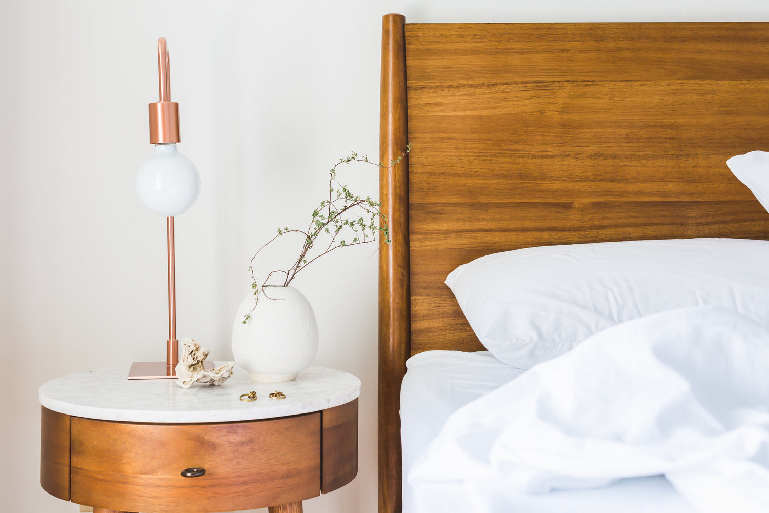 Mid-century modern bedroom with walnut wood headboard, round marble-top nightstand, copper table lamp, and white ceramic vase with delicate branches