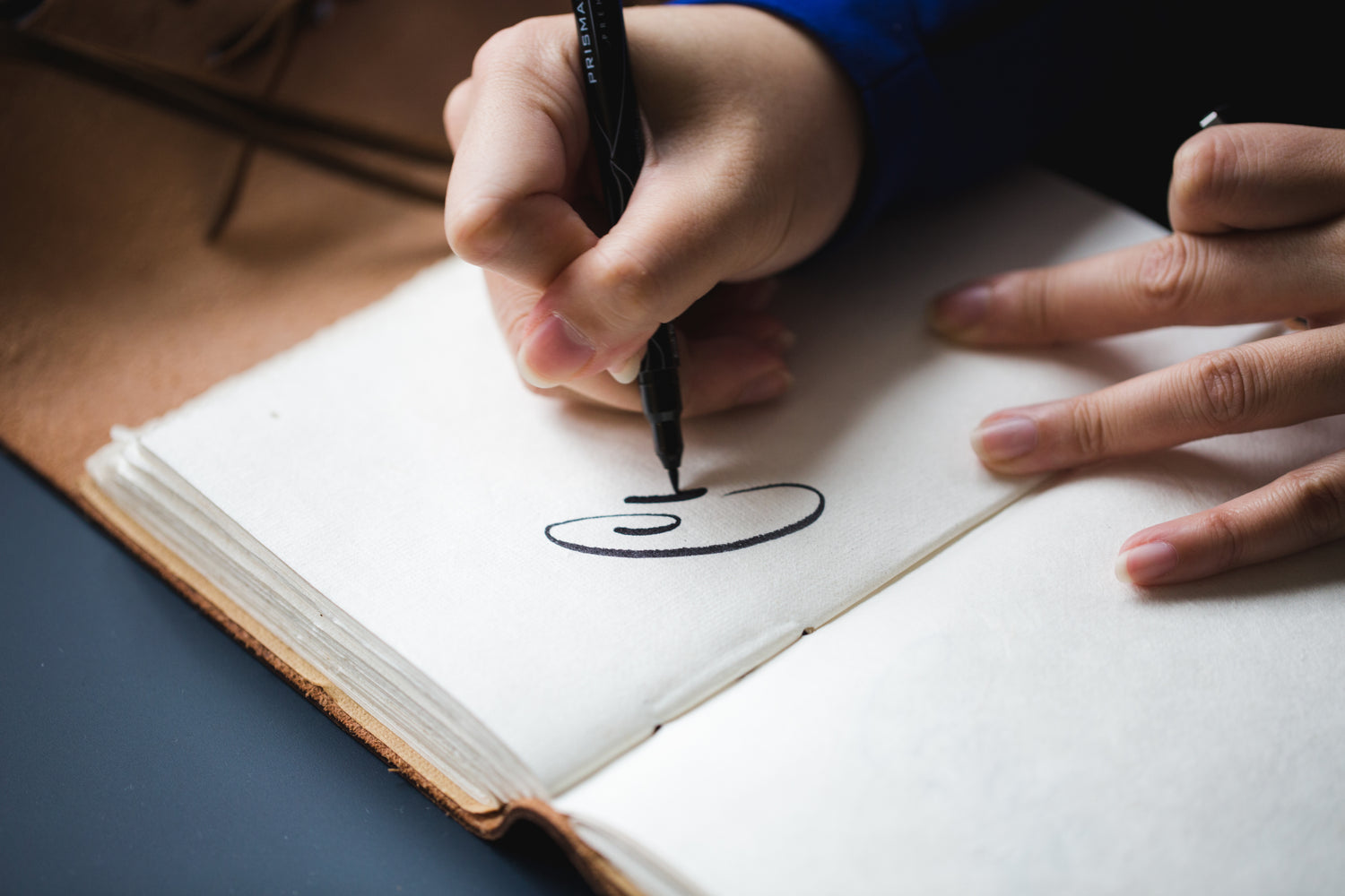 Close-up of a person’s hand using a black pen to draw a simple curved, abstract symbol in an open notebook, with the other hand resting nearby on the page.
